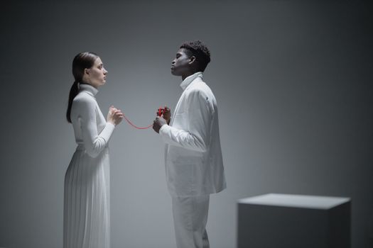 A couple dressed in white connected by a red thread in a minimalist studio setting.