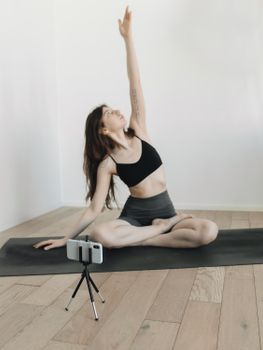 A young woman practices yoga indoors, using a smartphone on a tripod to record her session on a mat.