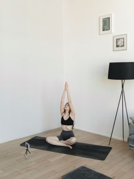 A woman in sportswear sitting on a yoga mat indoors, practicing a yoga pose while recording herself with a smartphone.
