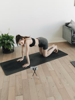 A woman performs a yoga pose on a mat indoors, with a phone recording her session.