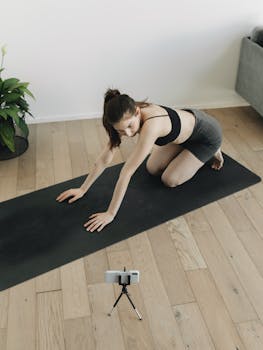 Young woman practicing yoga indoors on a mat, using smartphone for guidance. Focus on fitness and wellness.