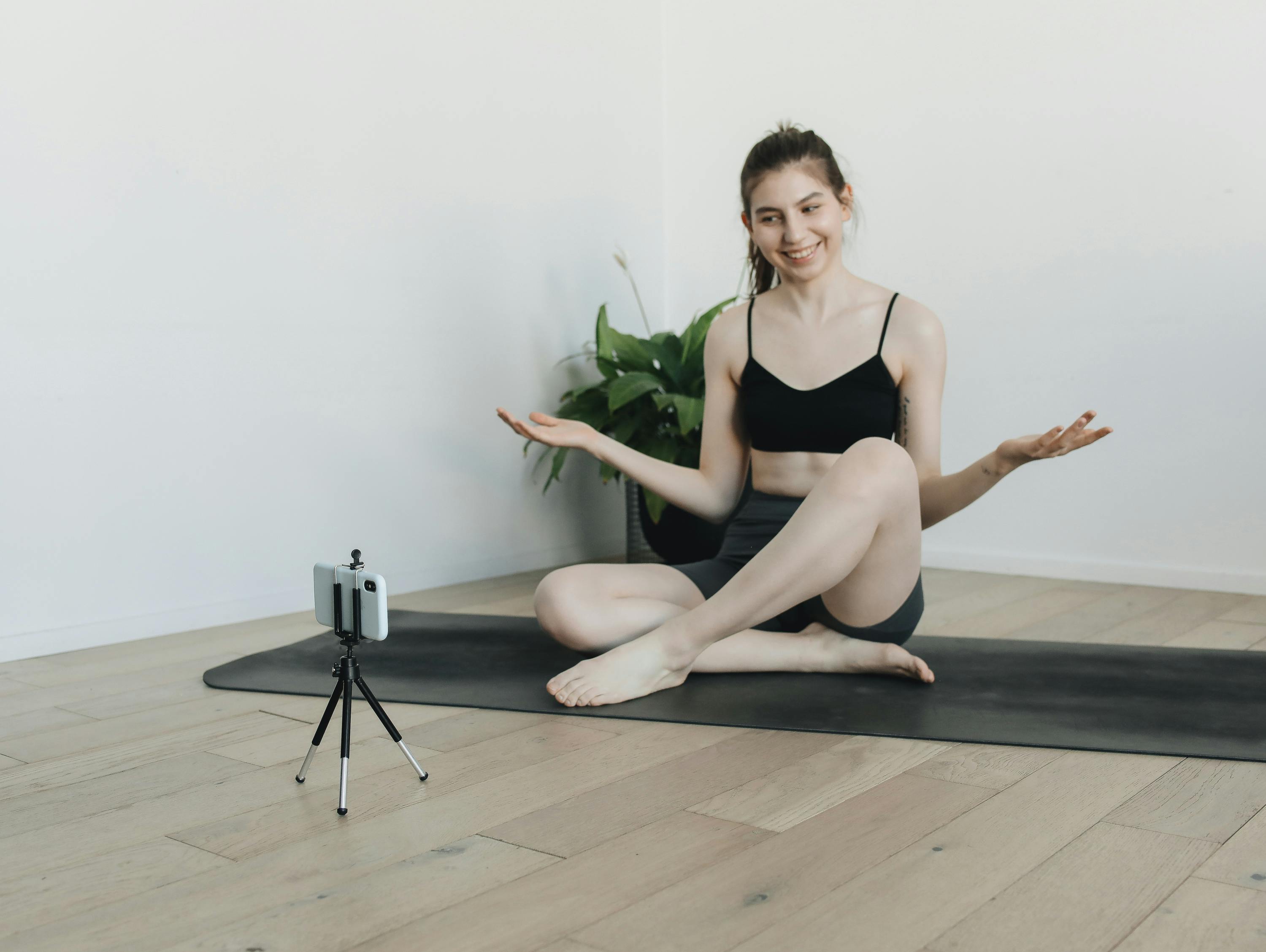 Smiling woman doing yoga on mat indoors with smartphone setup for vlogging.