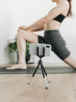 A woman doing yoga exercises on a mat with a smartphone setup for filming at home.
