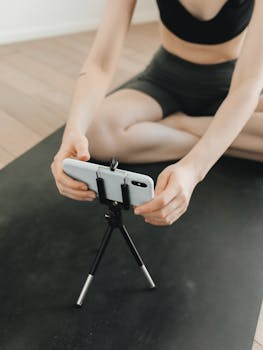 A woman unrecognizable settings up a smartphone on a tripod while preparing for a yoga session indoors.