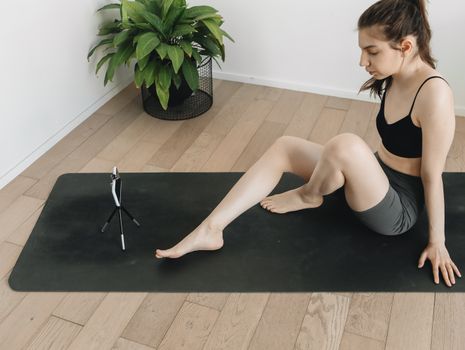 Young woman in activewear sitting on a yoga mat recording herself indoors. Ideal for fitness and lifestyle content.