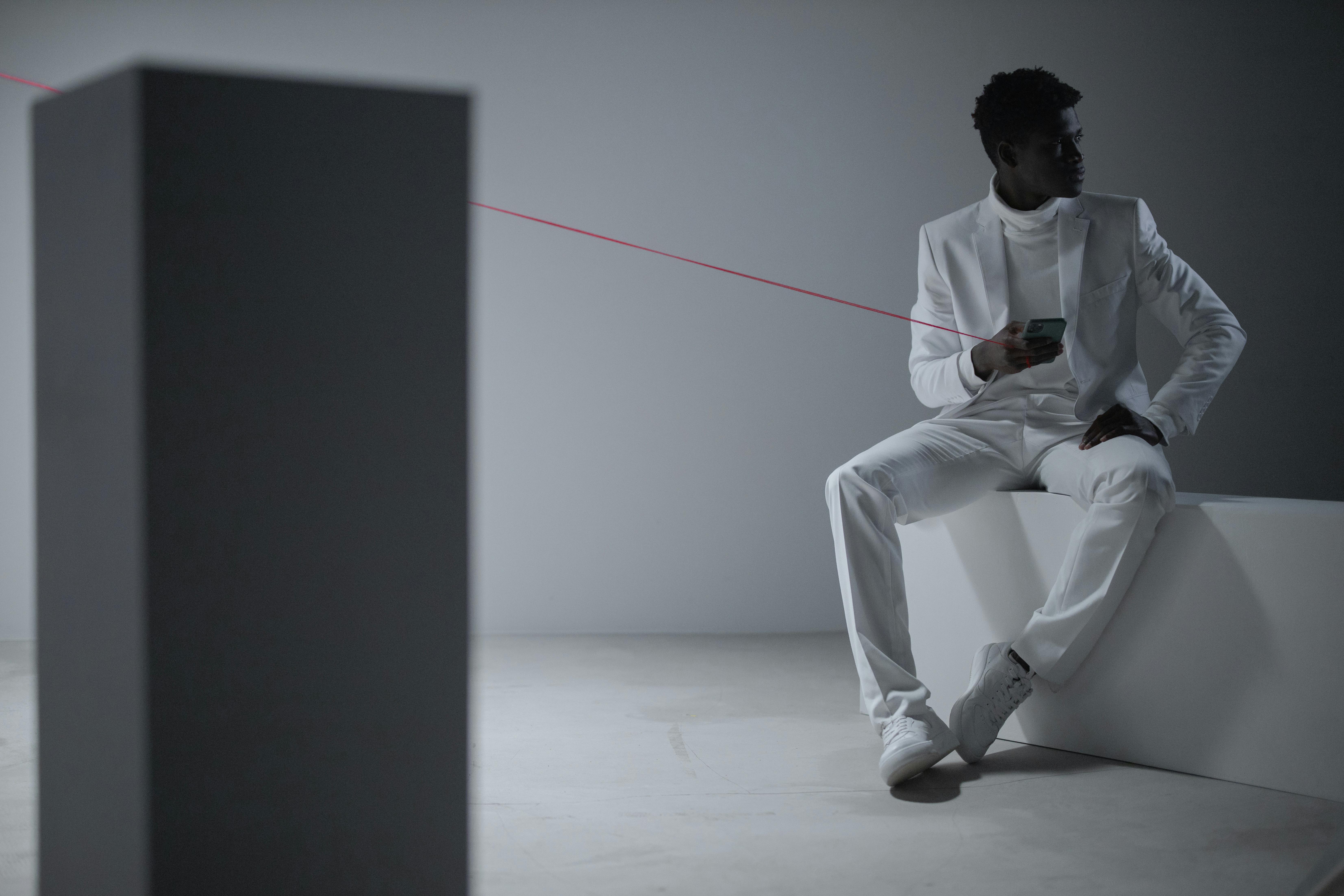 Stylish man in white suit with smartphone, sitting in a minimalist setting. Conceptual and modern fashion imagery.