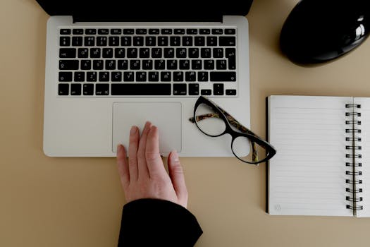 Hand on a laptop trackpad with notebook, eyeglasses, on a beige desk. Perfect for business or technology themes.