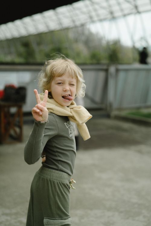 Free Blonde child in playful pose with peace sign, smiling outdoors with blurred background. Stock Photo