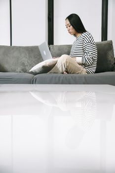 Asian woman sitting on a sofa, working remotely with a laptop at home, embracing digital nomad lifestyle.
