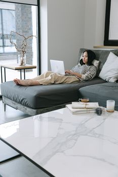 Asian woman lying on a sofa, focused on remote work using a laptop in a modern living room.