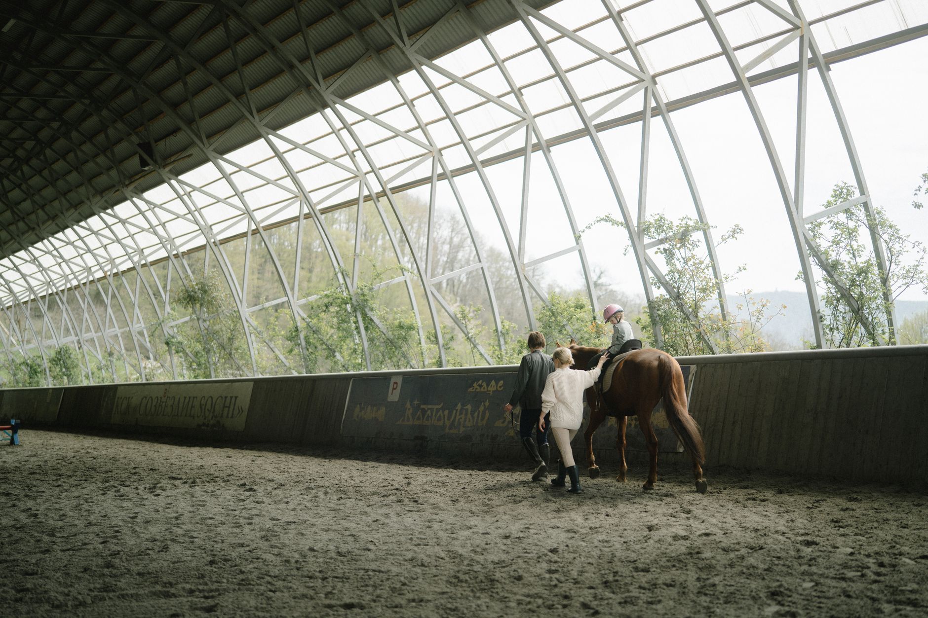 Child learning horseback riding in an indoor equestrian arena with guidance from instructors.