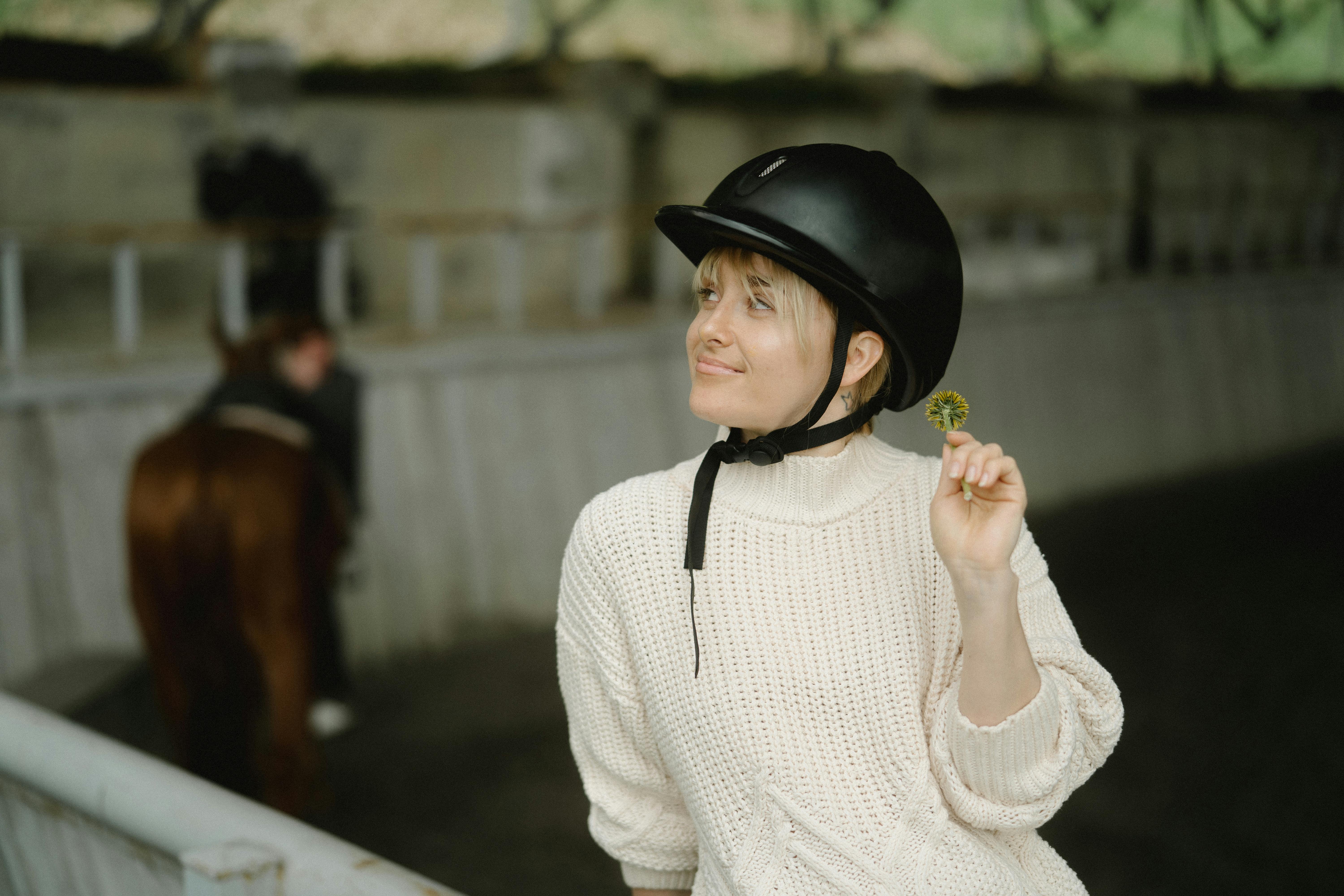 Woman wearing knit sweater and helmet holds a flower, enjoying the indoor equestrian setting.