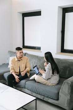 Two adults discussing work on a couch at home with a tablet and laptop.