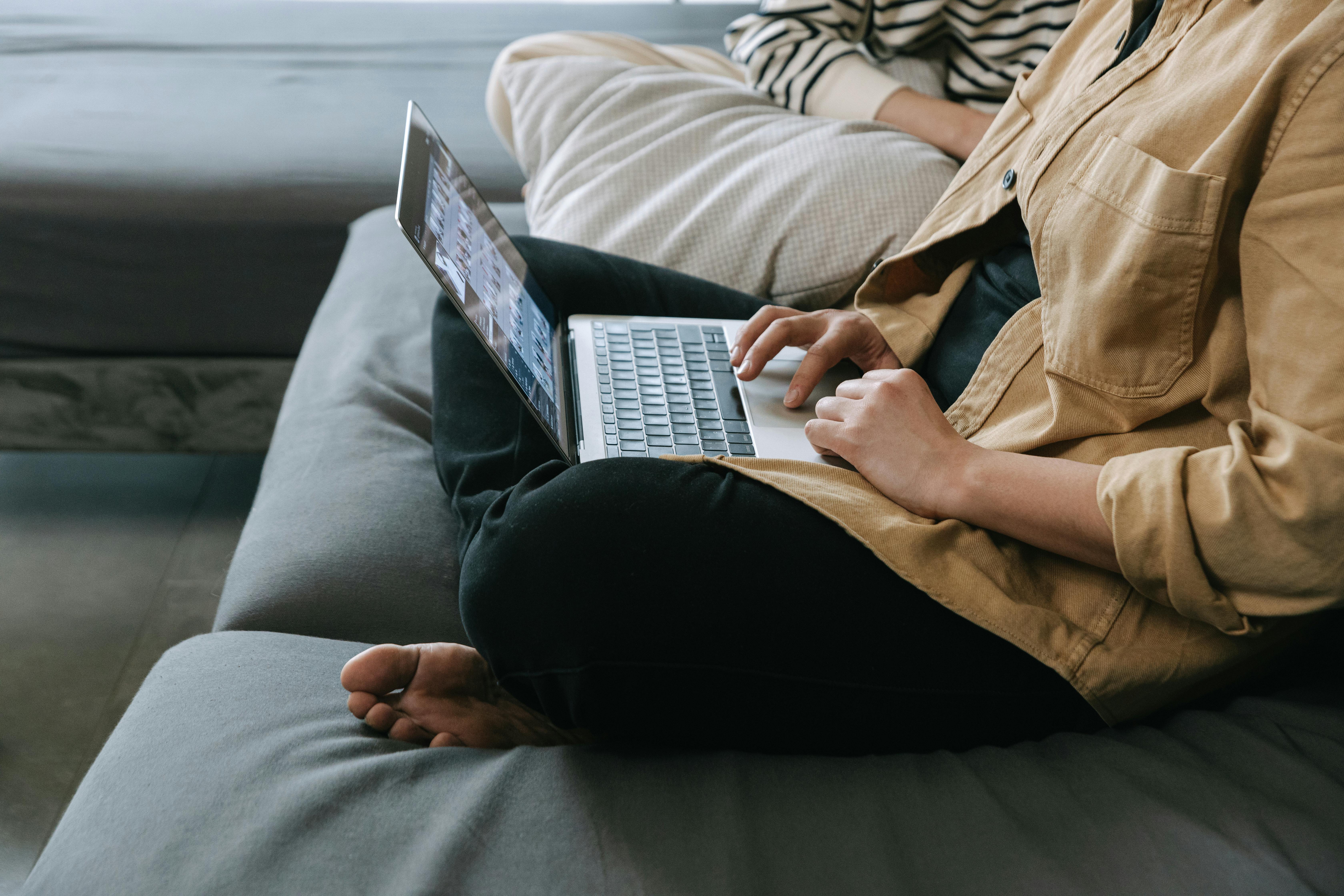 A person working on a laptop while sitting cross-legged on a comfortable couch indoors.