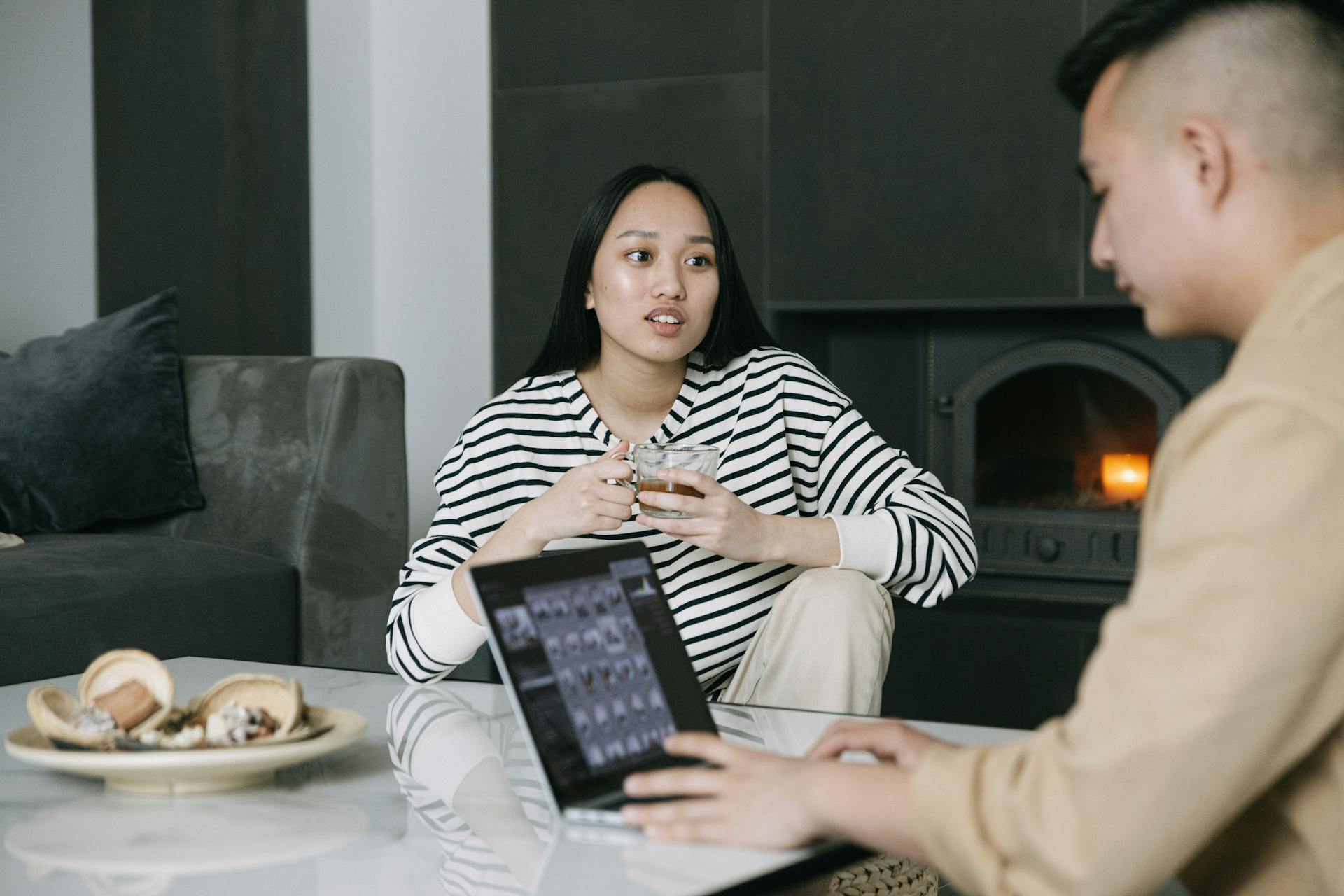 Asian couple discussing while drinking coffee and using a laptop near a cozy indoor fireplace.