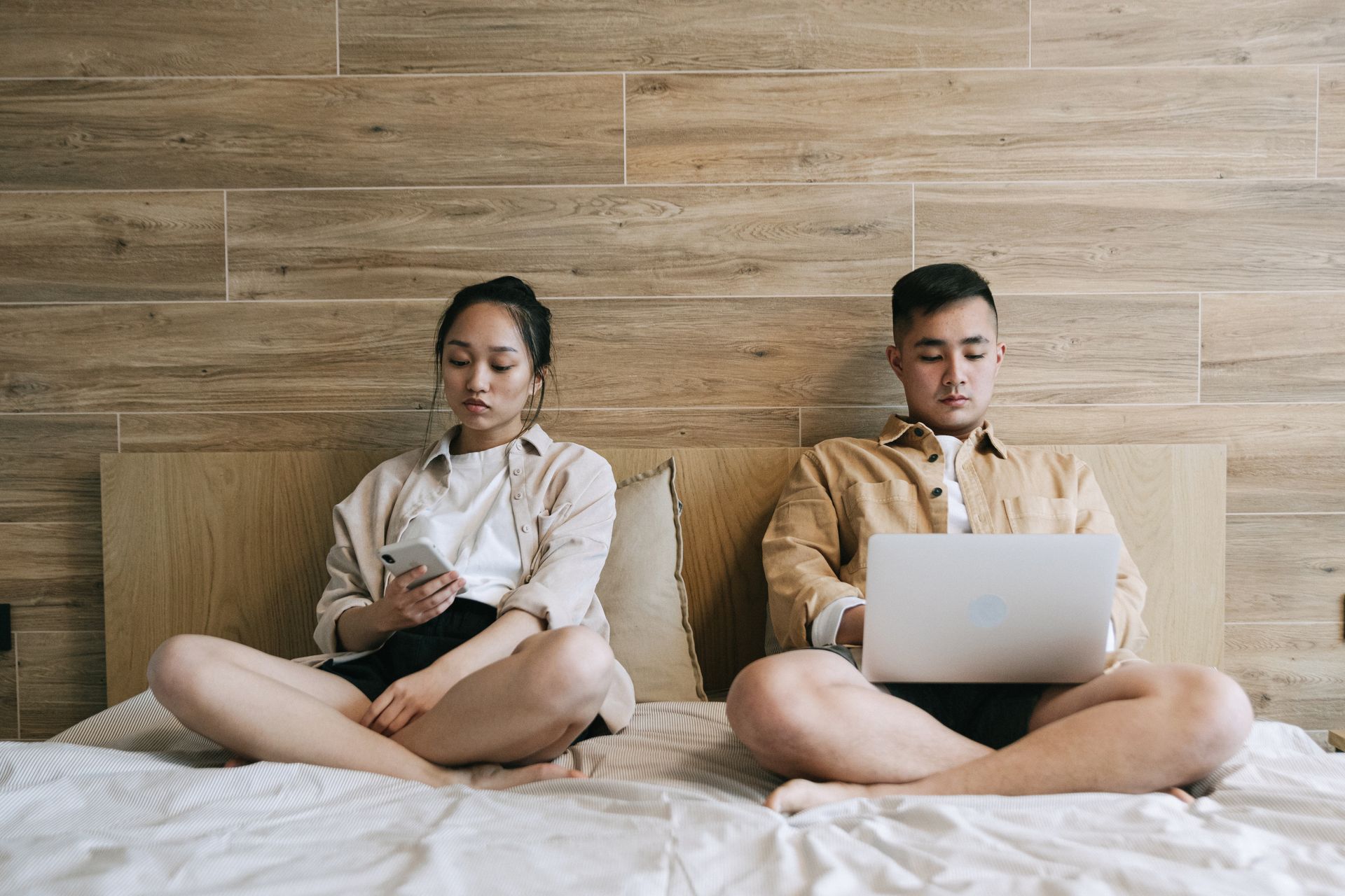 Young couple sitting on a bed, using smartphones and laptops in a modern bedroom setting.