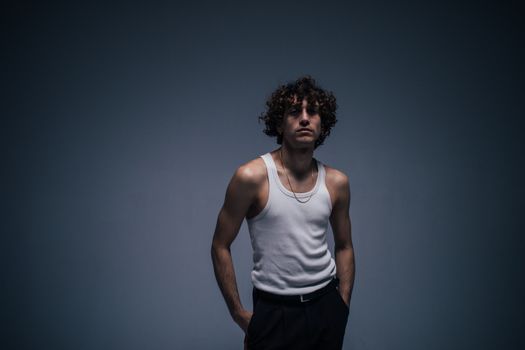 Portrait of a young man with curly hair in a white tank top posing in a moody studio setting.