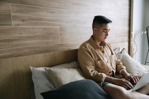 Adult male using a laptop while sitting on a bed in a cozy bedroom setting.