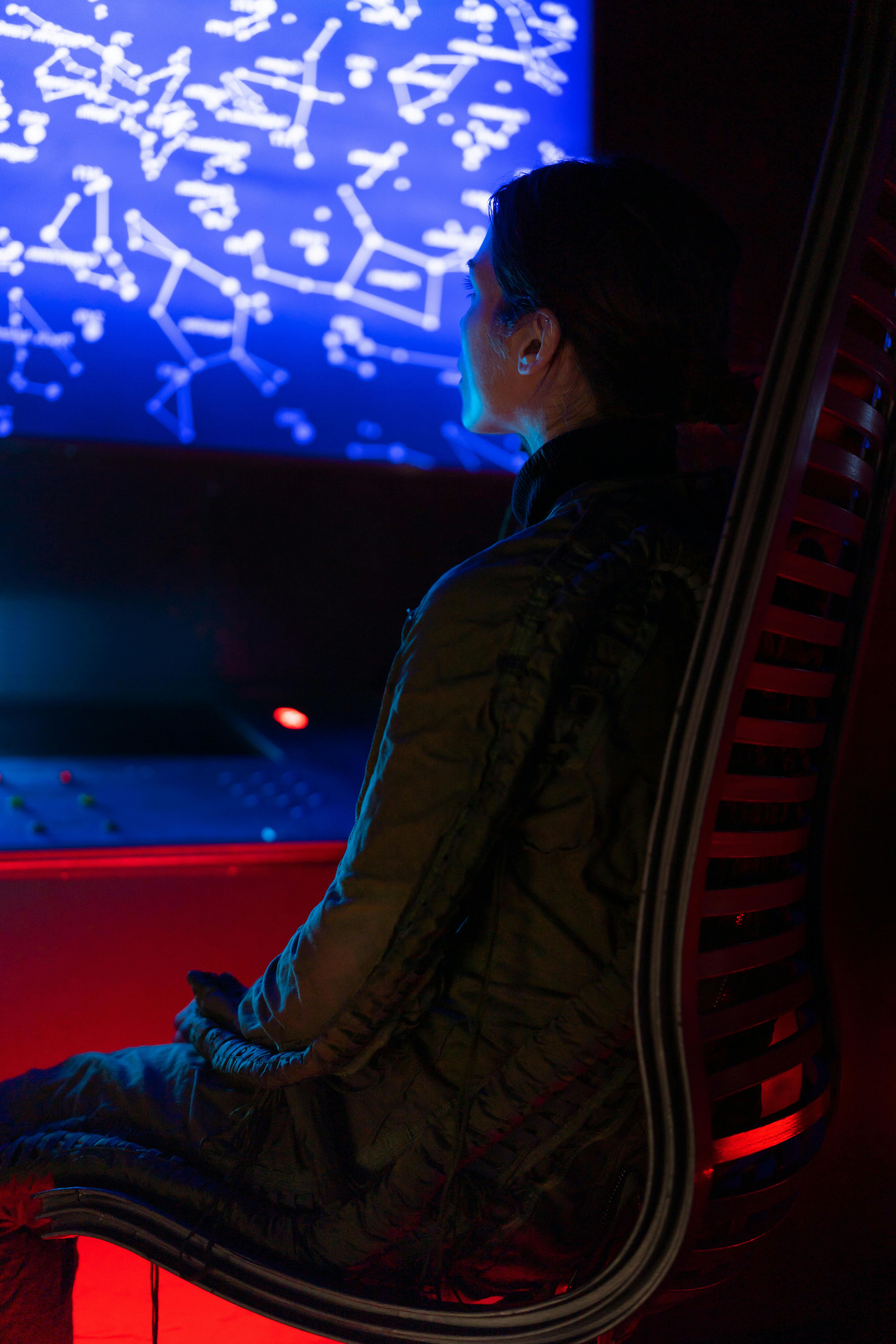 A woman in a space suit observes a high-tech control panel displaying a galaxy map.