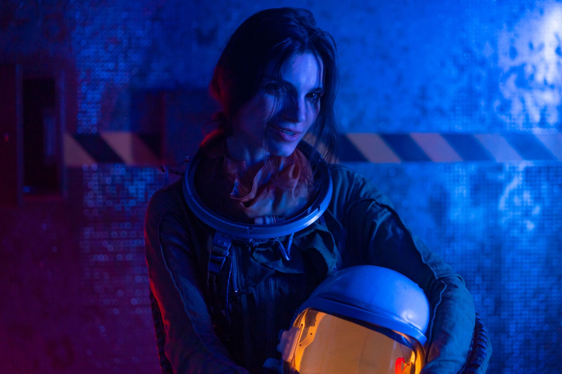 A dramatic portrait of a female astronaut holding a helmet in a creatively lit indoor setting.