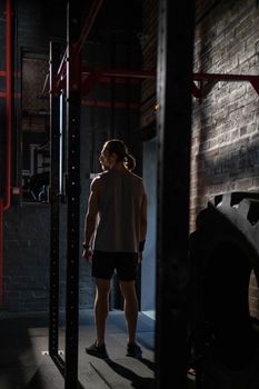A muscular man stands in a dimly lit gym, wearing sportswear, exuding fitness and strength.