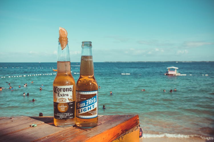 Two Corona Extra And San Mig Light Beers On Top Of Brown Wooden Plank Near Beach