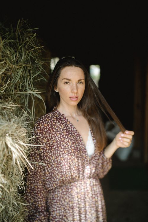 Free An adult woman in a floral dress poses indoors next to hay, bathed in natural light. Stock Photo