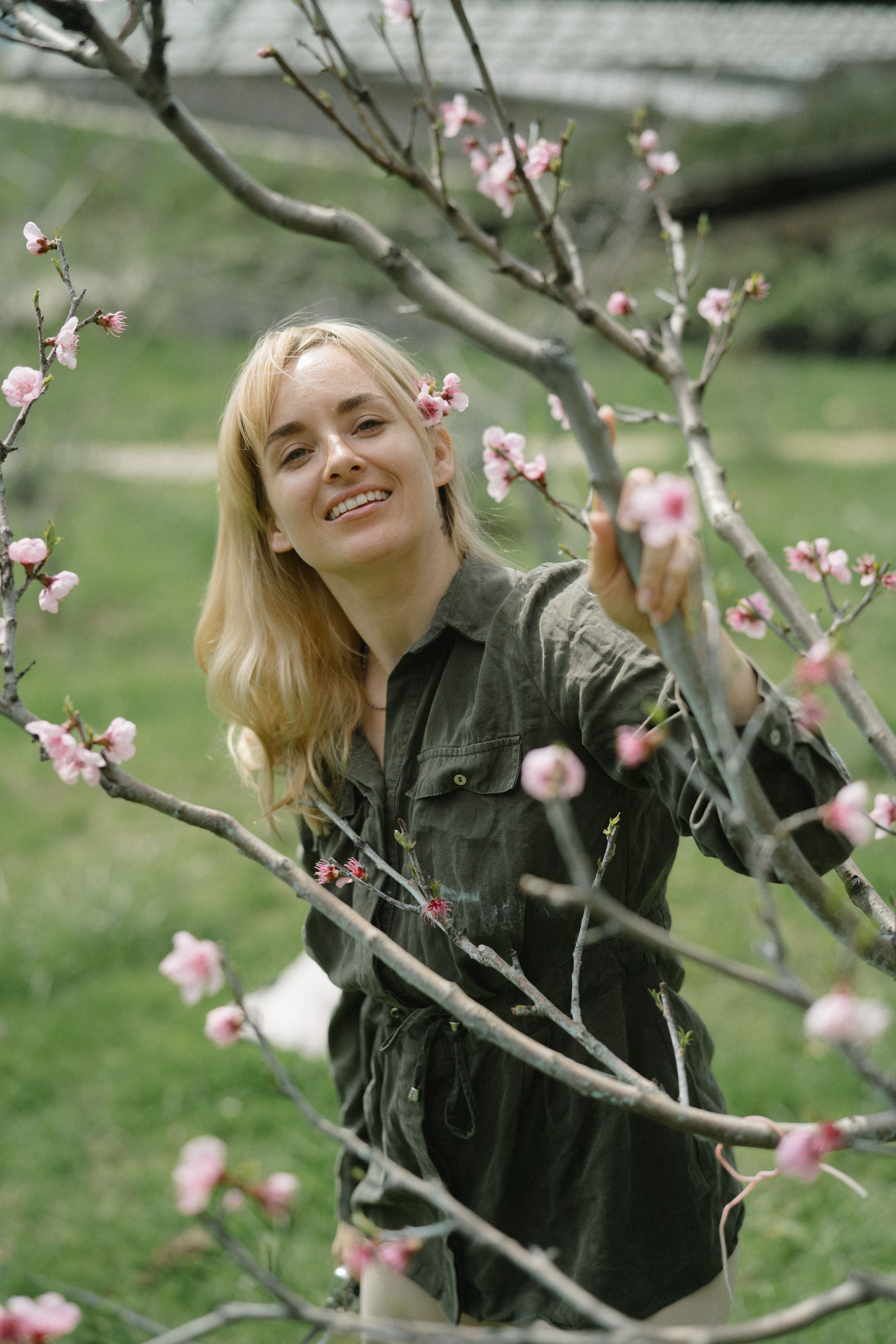 A woman smiling while standing among cherry blossoms in a sunny spring garden.