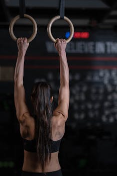 Back view of a woman exercising on gymnastic rings in a dimly lit gym setting, showcasing strength.