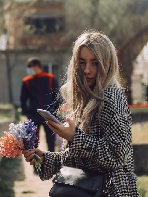 Free Blonde Woman Using Her Cell Phone while Holding a Bunch of Flowers Stock Photo