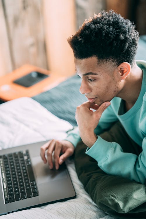 Side view of concentrated African American male freelancer in casual clothes surfing modern netbook while lying on bed at home