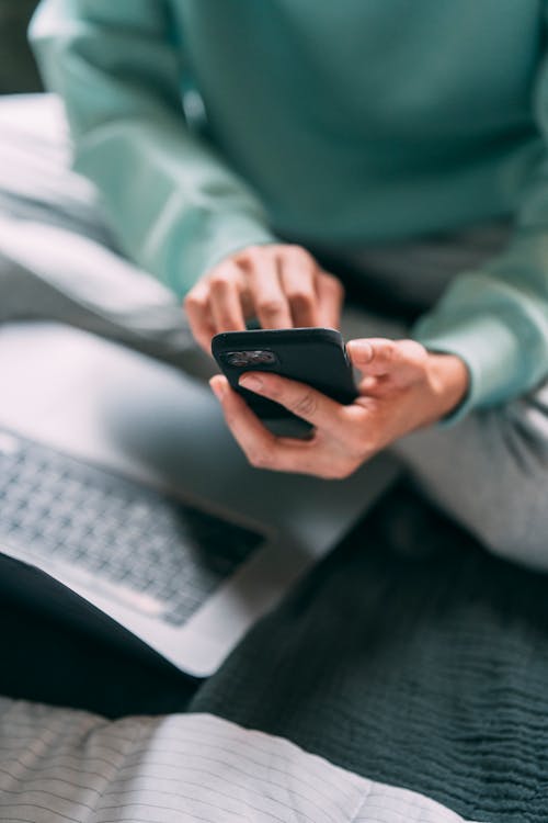 Free Crop anonymous male freelancer text messaging on modern cellphone while sitting on bed with laptop during remote work at home Stock Photo