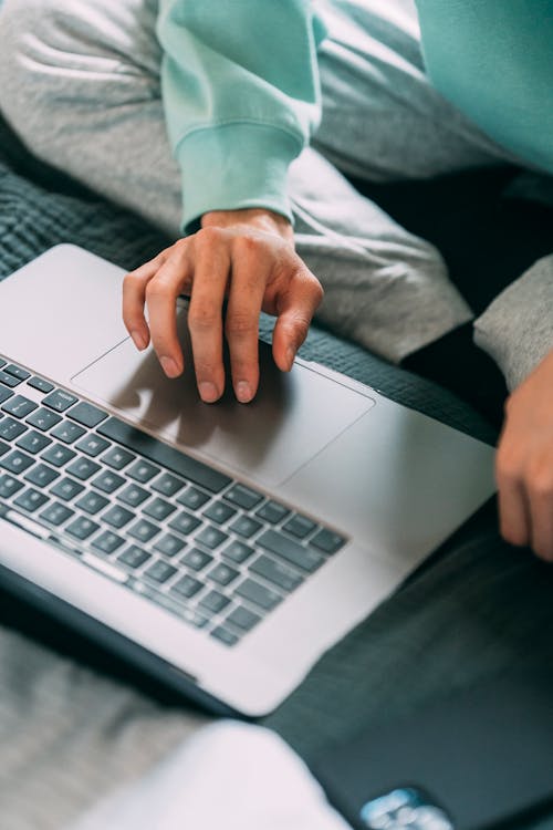 Crop faceless male freelancer browsing internet on netbook while sitting on comfortable bed during online work in bedroom at home