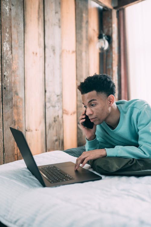 Free Side view of concentrated African American male freelancer browsing netbook while having phone call on bed during remote work at home Stock Photo
