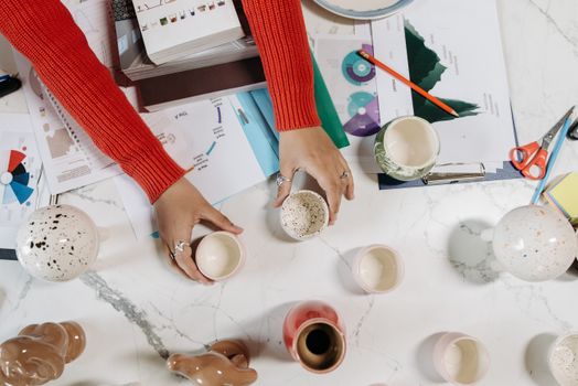 Hands arranging ceramic cups on a marble table with charts and notebooks, perfect for creative workspace themes.