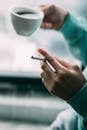 Crop anonymous male smoker with cigarette in hand drinking hot coffee from cup while sitting near window in light room