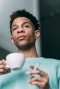 Low angle of serious African American male with cup of coffee and cigarette in hand standing in room at home