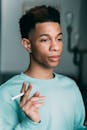 Pensive African American male smoker with unlit cigarette in hand looking into distance while standing in light room at home