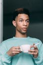 Pensive African American male smoker with white cup of coffee and cigarette in hand standing in light room at home