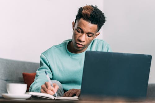 Contemplative black man with dyed hair taking notes near laptop