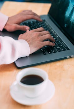 From above of crop anonymous male typing on laptop while sitting at wooden table with cup of coffee