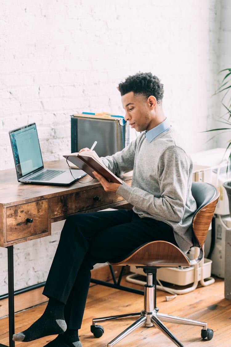 Black Man Writing In Planner At Table With Laptop