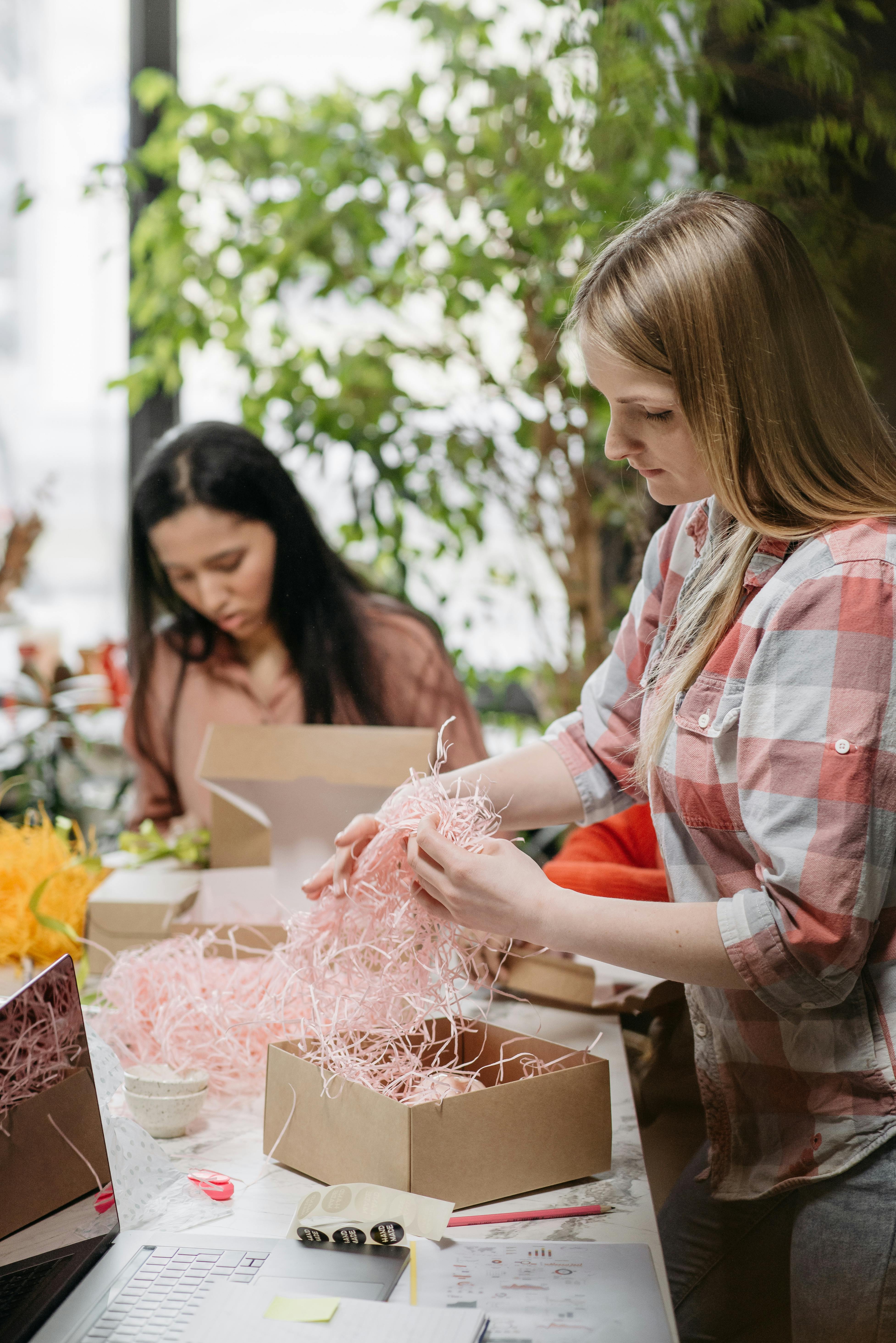 Two women arranging shredded paper into gift boxes indoors, teamwork concept.