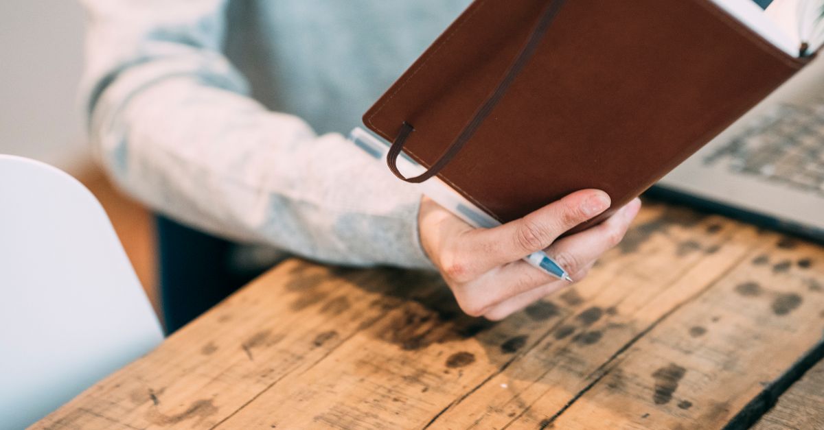 Crop unrecognizable male examining brown planner at timber table on blurred background in office