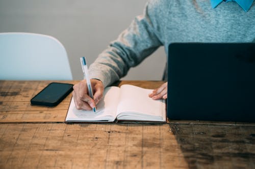 Crop faceless male writing in notebook at wooden table with netbook and blank smartphone on gray background