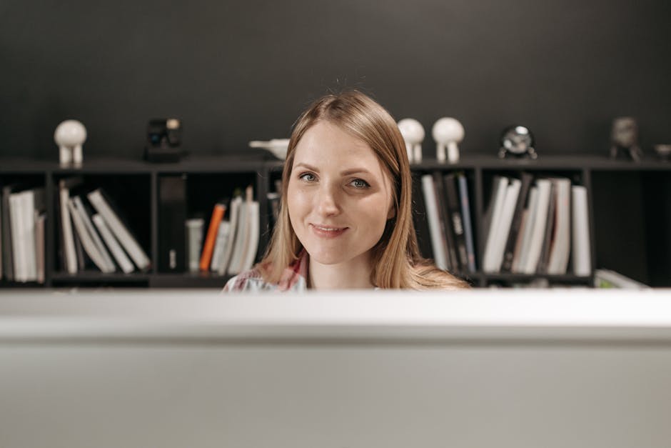 Friendly young woman at her desk with bookshelves in modern office setting.
