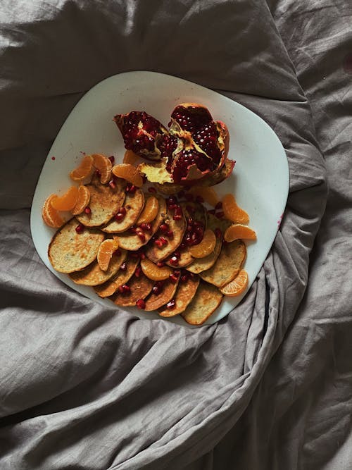 Top view of delicious homemade pancakes with fresh ripe pomegranate and tangerine slices served on white ceramic plate placed on comfy bed