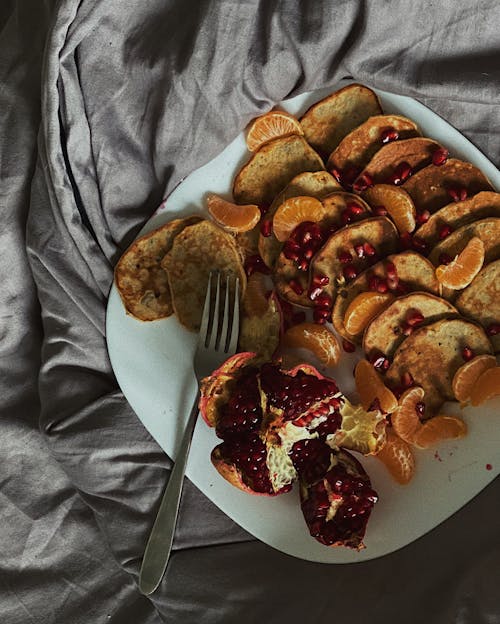 Fresh mandarin slices and pomegranate served on plate with pancakes on bed