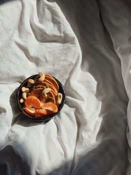 From above of bowl with slices of healthy ripe oranges with peanuts and yummy pancakes placed on comfy bed in sunny morning
