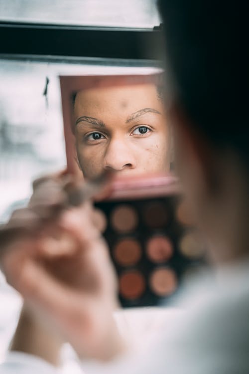 Free Crop blurred feminine African American male with palette of colorful eyeshadows looking at mirror while applying makeup in light room Stock Photo
